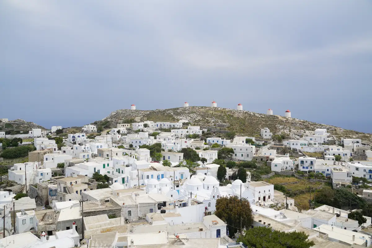 A hillside of flat-roofed white houses and small domed churches packed together, with a bare ridge above them carrying a line of round stone windmills with red caps.