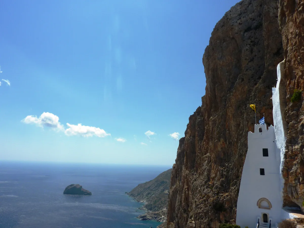 A narrow white building pressed into a hollow in a sheer brown cliff face high above the open sea, flags flying from its roof, with a small islet in the water far below.