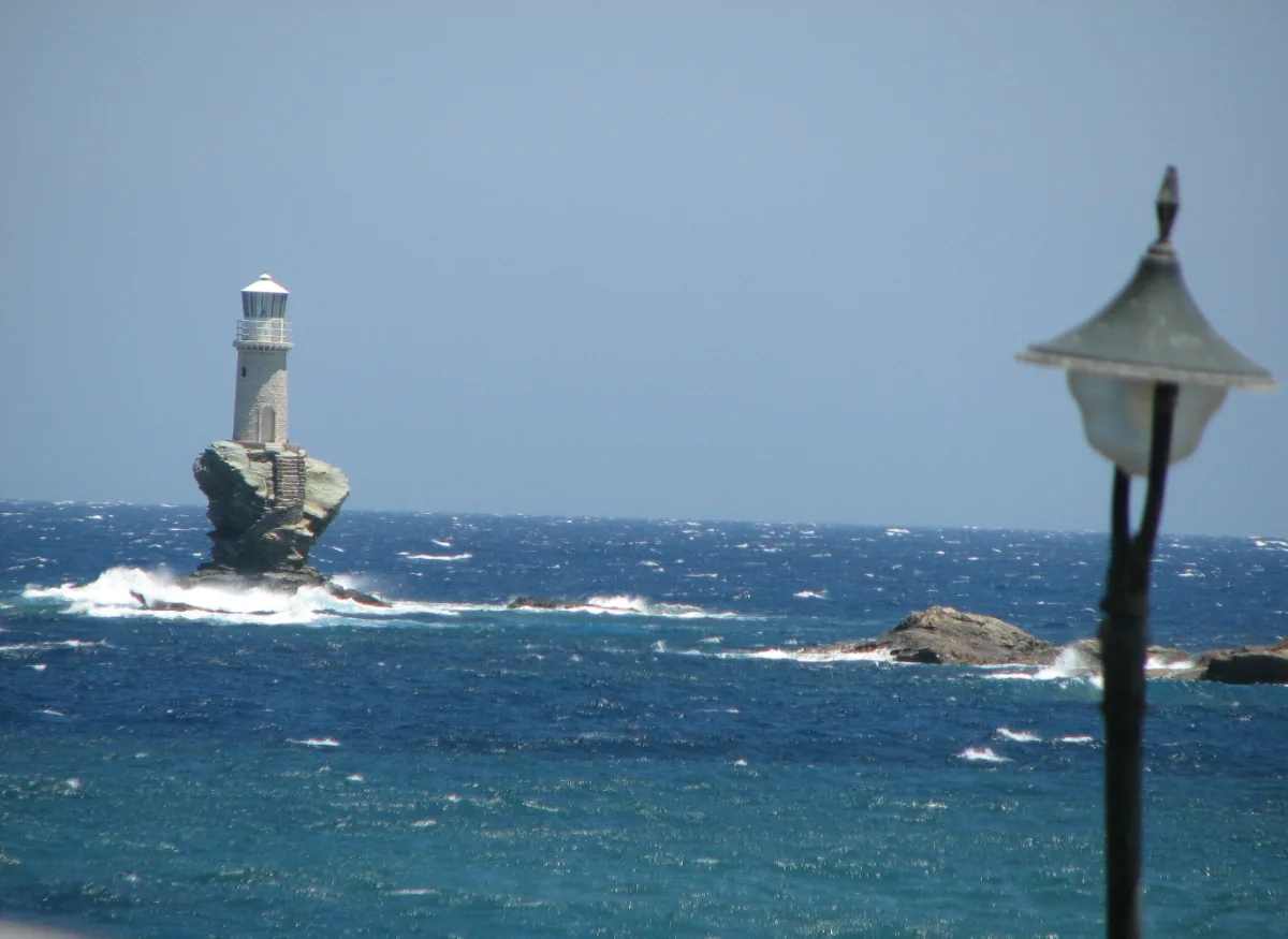 A slim stone lighthouse with a white lantern room standing on the flat top of an isolated rock stack in open sea, with steps cut into the rock running up to its door and waves breaking on low rocks beside it.