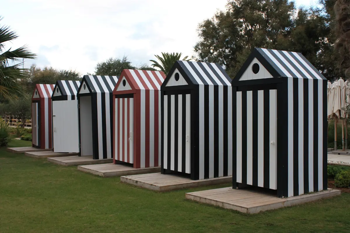 A row of striped wooden changing huts on a lawn at Astir Beach on the Athens Riviera, with furled parasols stacked behind them