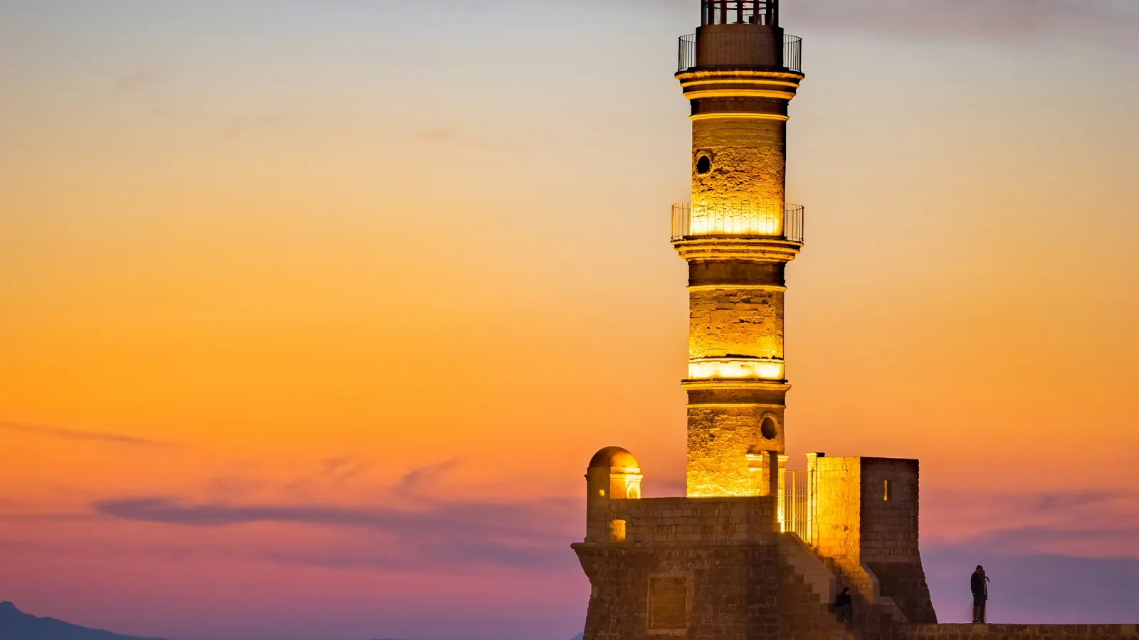 The lighthouse at the mouth of the Venetian harbour of Chania at sunset: the tall slender stone tower stepping inward through three tapered stages with railed galleries and a lantern on top, standing on a stone bastion at the end of the rubble breakwater