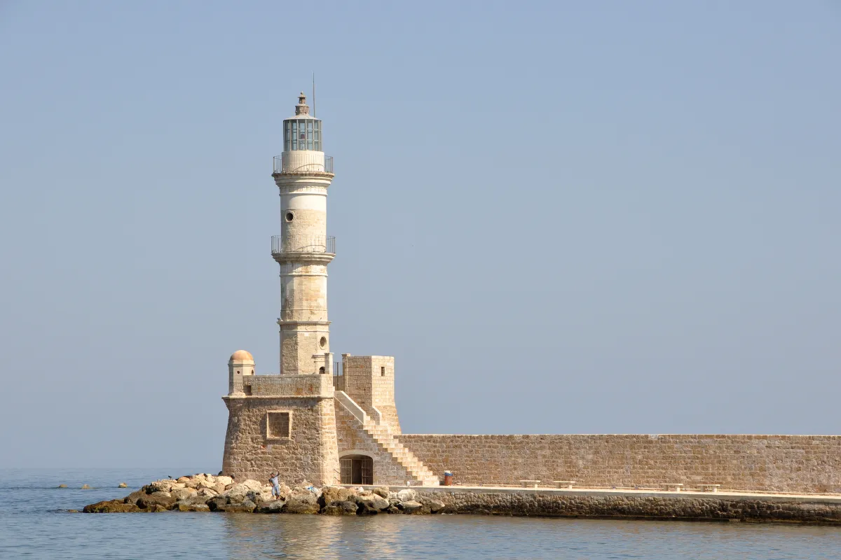 A tall stone lighthouse standing at the end of a low stone breakwater, with calm sea in front of it and the hills behind the town in the distance.