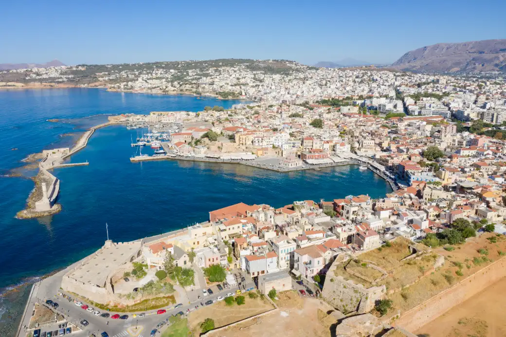 An aerial view of a curved harbour basin ringed by a quay, with a long stone breakwater closing the mouth and a dense old town of tiled roofs packed behind it.