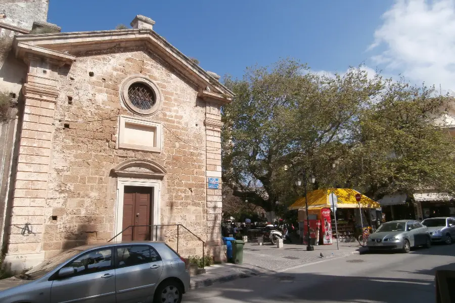 A small stone church with an arched doorway and a round window standing on the edge of an old-town square, with plain buildings and a street running off to one side.