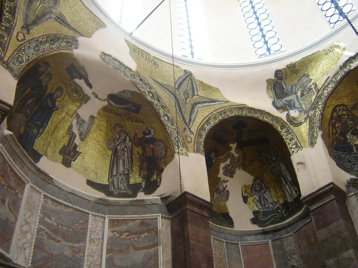 Gold-ground mosaics filling the curved recesses beneath a church dome: standing saints and figured scenes set against fields of gold, a six-winged seraph at the centre, Greek lettering in the gold above the figures, and bare plaster where tesserae have been lost.