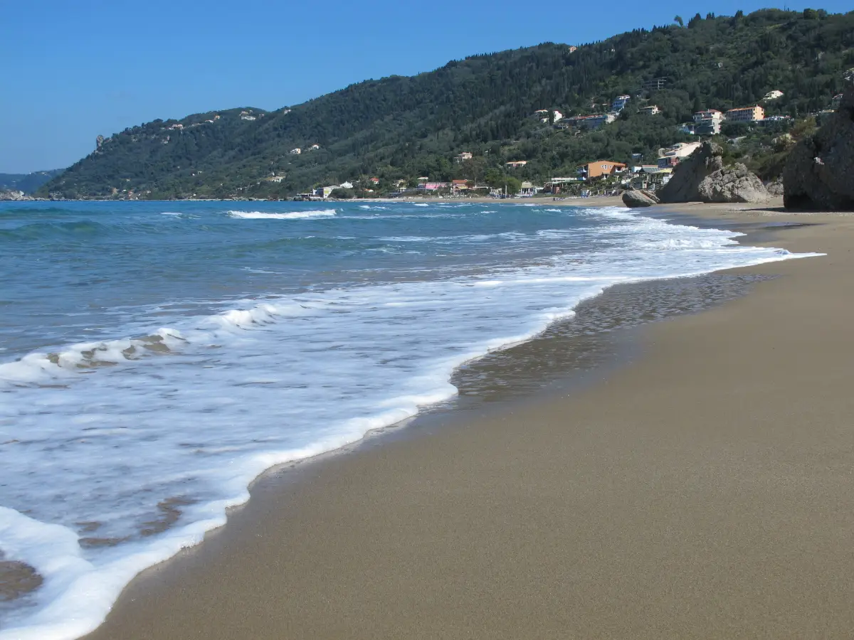 Surf running up the sand at Agios Gordios on the west coast of Corfu, seen from the waterline, with the village scattered over the green hillside behind and a headland closing the far end