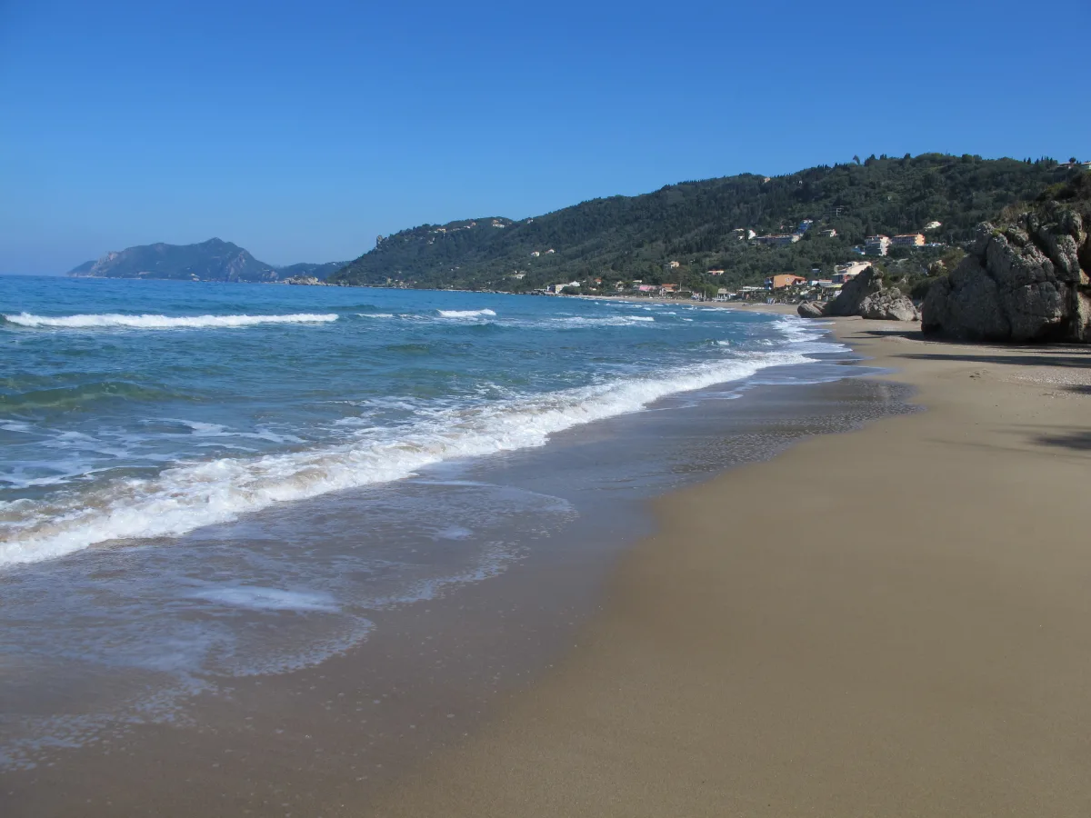 Surf running up the empty sand at Agios Gordios on the west coast of Corfu, with the village on the green hillside behind and a headland closing the far end of the beach