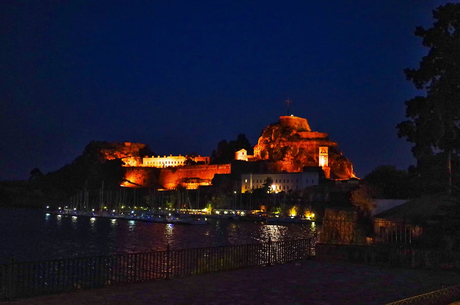 The Old Fortress of Corfu floodlit at blue hour above the marina, its clock tower lit and the water carrying its reflection