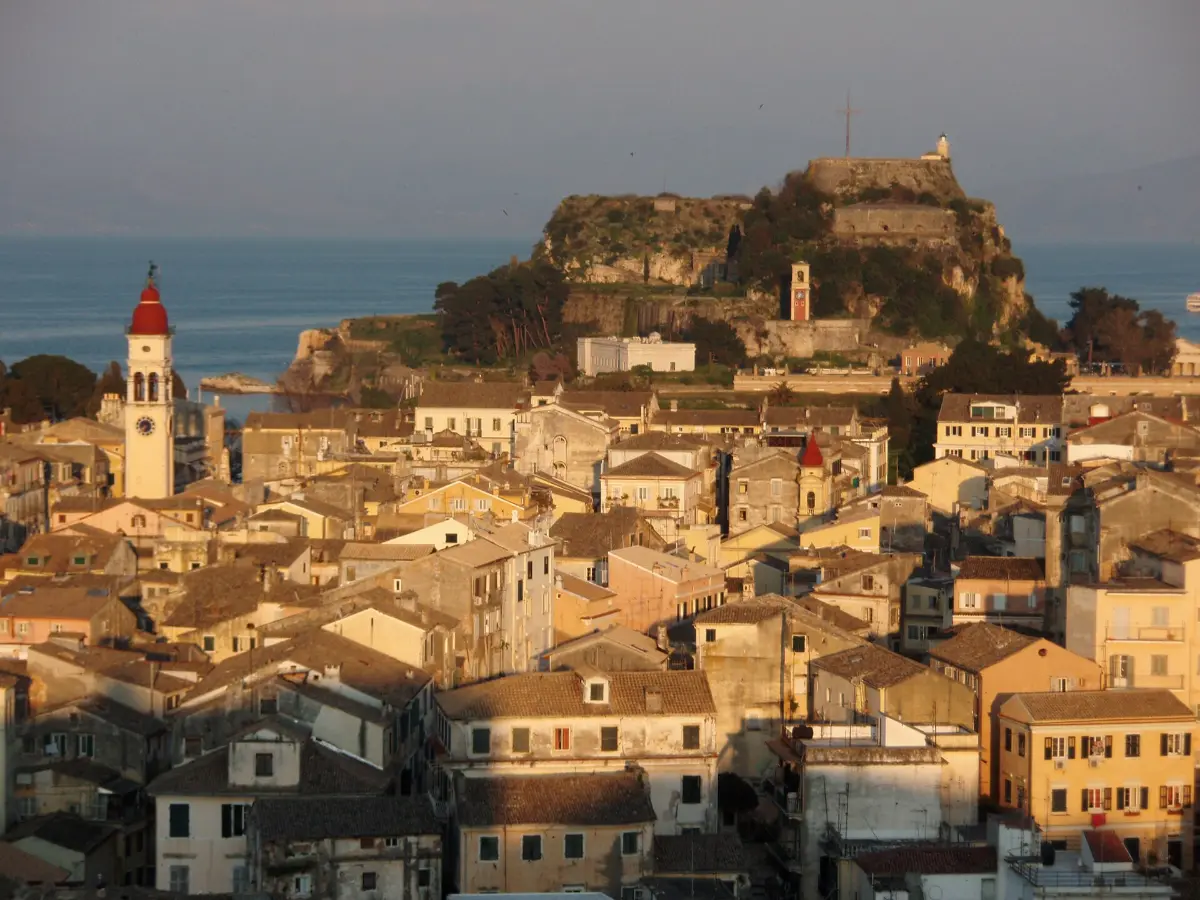 Corfu Old Town at sunset seen from above, tiled roofs and the red-domed bell tower of Agios Spyridon with the Old Fortress headland behind