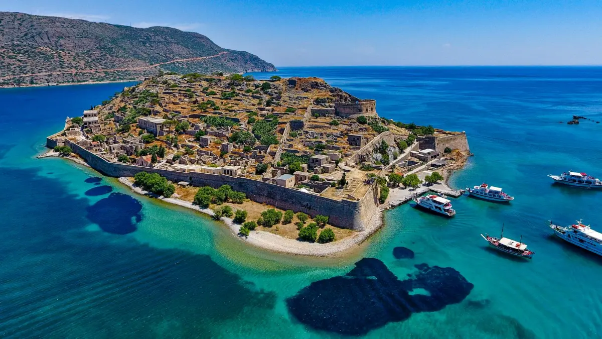 An aerial view of Spinalonga, a small fortified island off Crete - a Venetian wall running along its shore, the roofless ruins of the abandoned village on the slope, and tour boats moored alongside