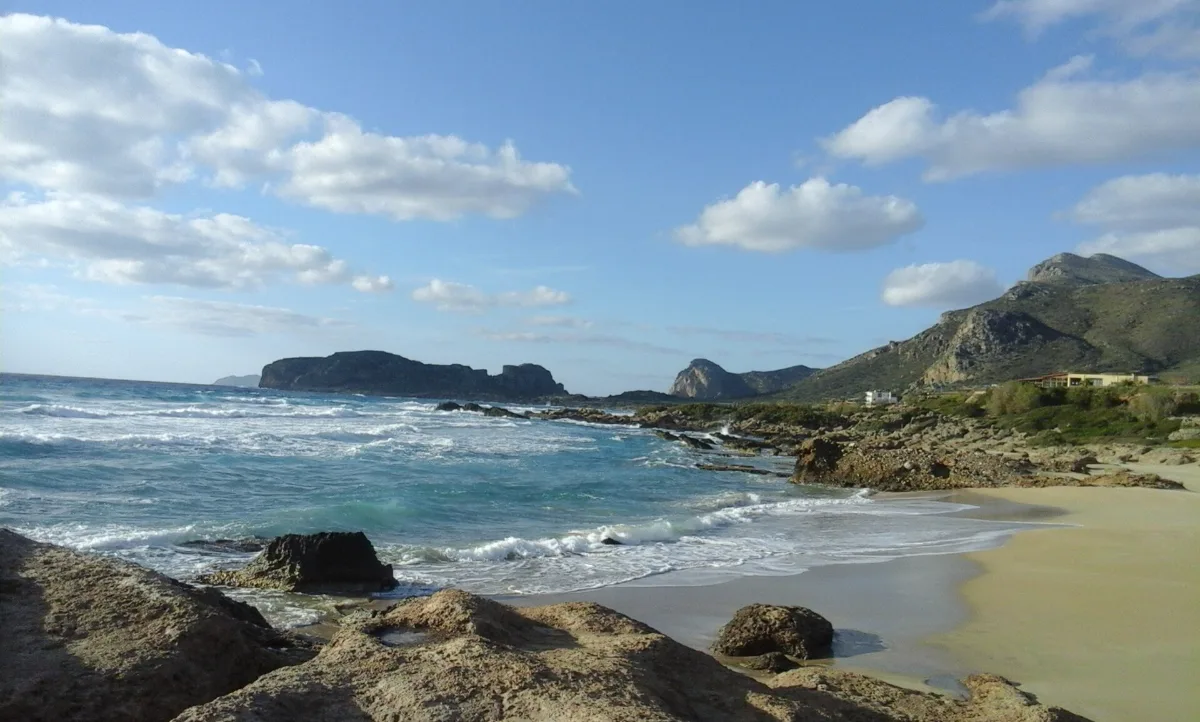 Waves rolling onto the wide pale sand of Falassarna beach on the exposed west coast of Crete, with a rocky headland behind