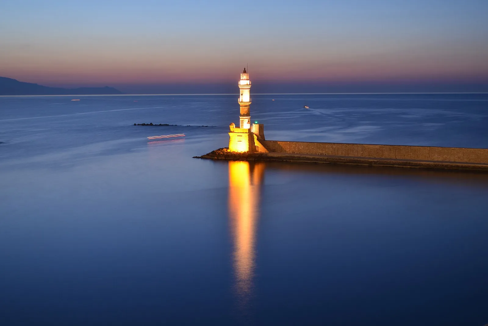 The Venetian lighthouse of Chania lit at blue hour at the end of the harbour mole, its reflection running across still water