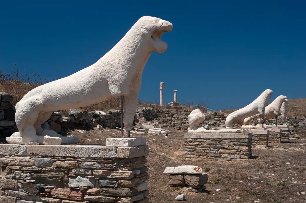 A row of lean white marble lions on stone plinths, mouths open and forelegs braced, standing along a low terrace with the stumps of two columns and dry hillside behind them.