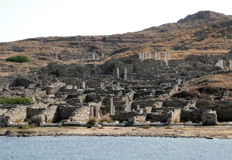 A dense field of ancient stone house walls, doorways and standing column shafts covering a low slope that runs down to the shoreline, with a bare brown hill above.