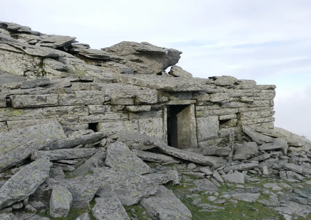 A low building of huge unmortared stone slabs on a bare rocky summit, with a squared doorway under one enormous lintel stone and a roof of overlapping flat slabs, loose rock scattered all around it.