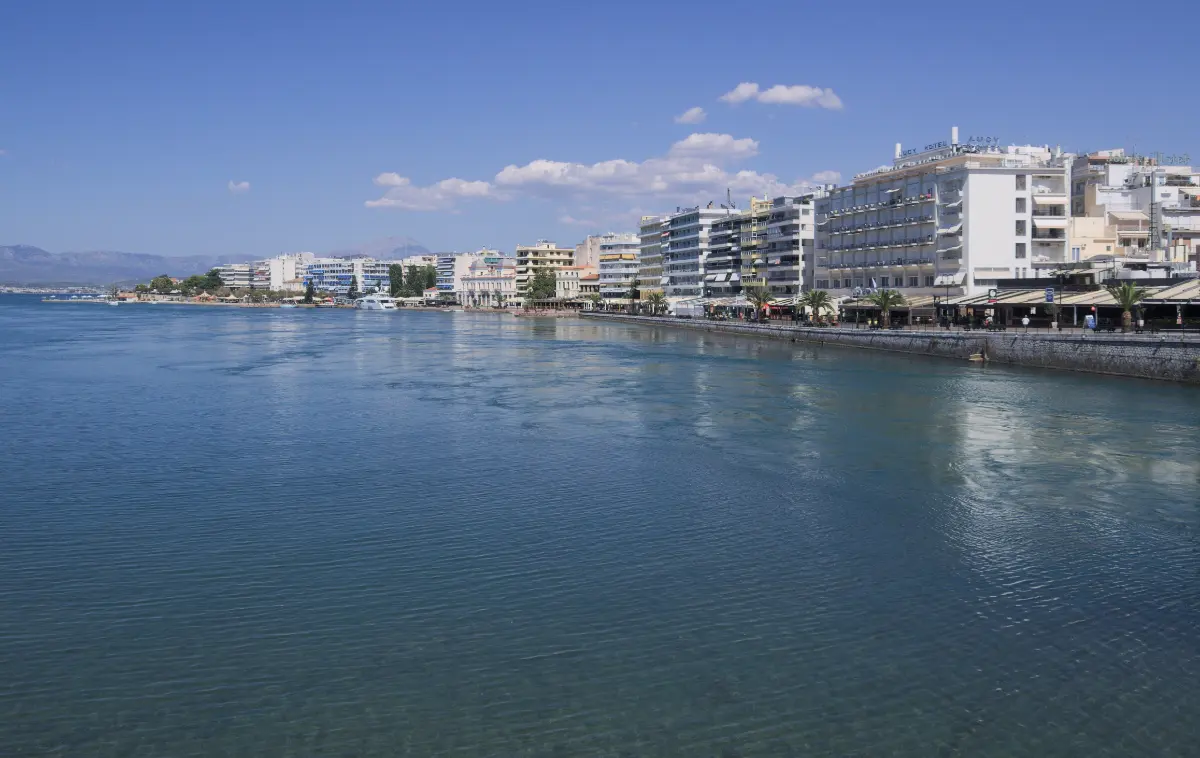 A wide channel of green water in the foreground, its surface streaked with pale swirls and slicks, with a town seafront of apartment blocks, hotels and a palm-lined promenade along the far bank.