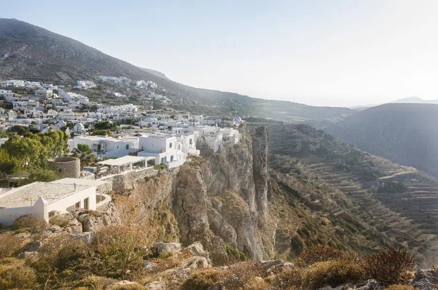 A whitewashed hillside town standing on the lip of a rock cliff that falls away in a sheer face, with bare terraced hills and hazy sea beyond it.