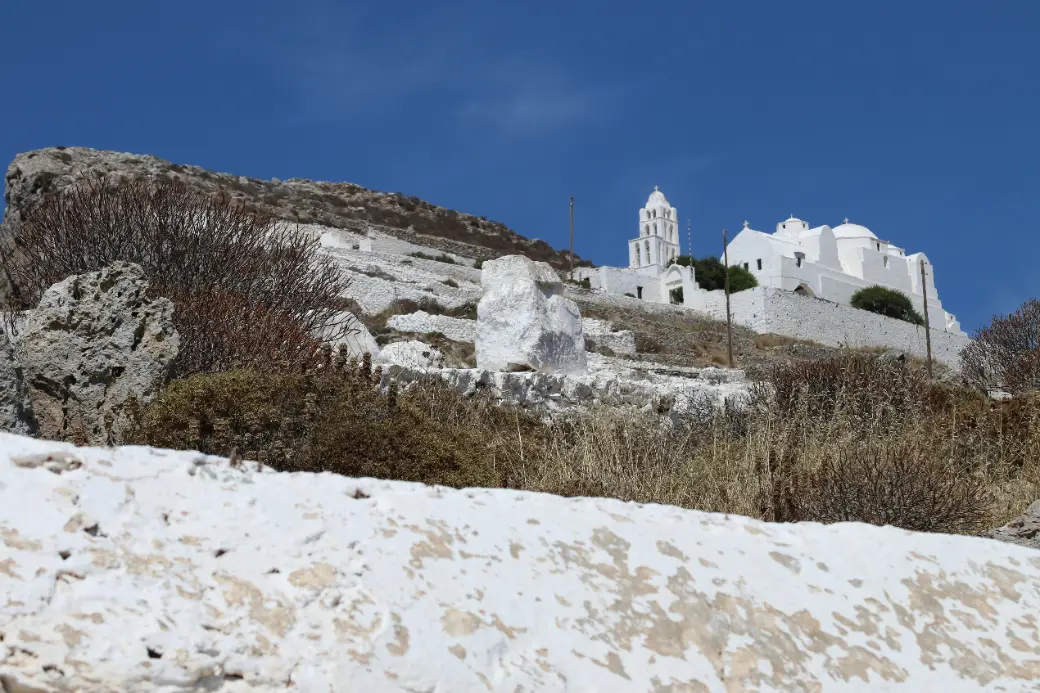 A white church with a low dome on a bare hilltop, seen from below, with a free-standing three-arched bell tower beside it and whitewashed retaining walls stepping up the rocky slope in front.