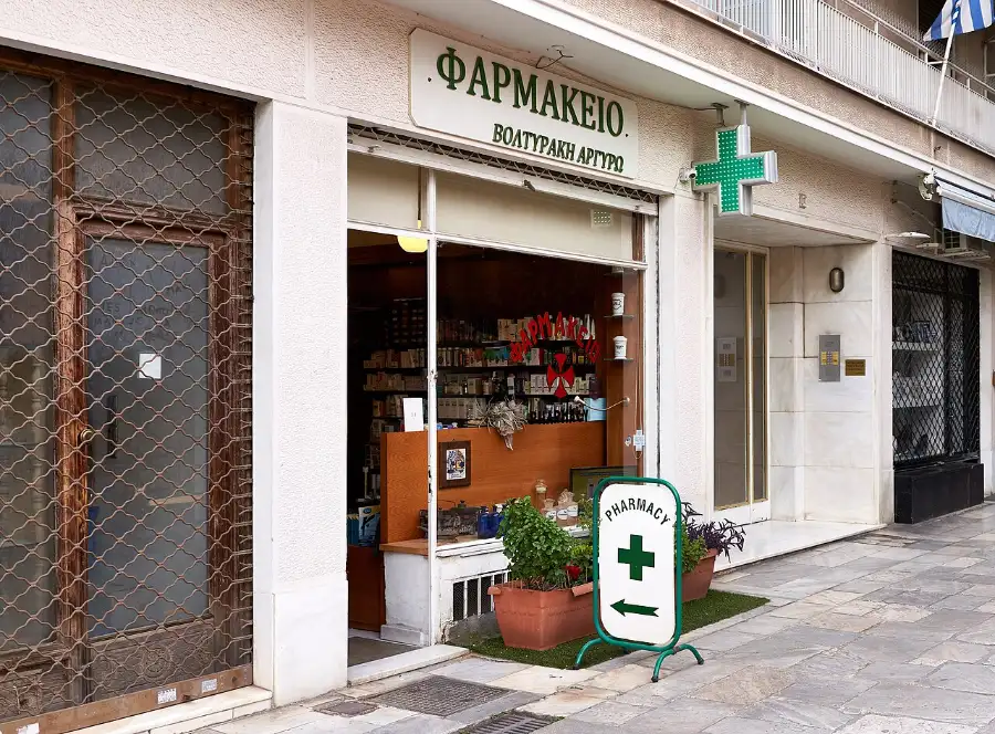 Illuminated green cross projecting over a pavement above a Greek pharmacy shopfront marked ΦΑΡΜΑΚΕΙΟ