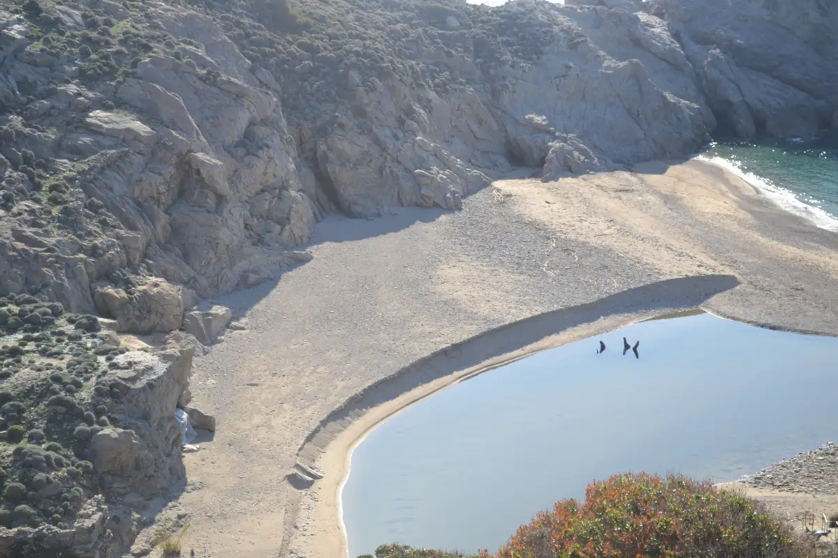 Looking down from a cliff onto a small cove: a bank of grey shingle separating a still, pale river pool from the open sea, with bare rock walls closing in on both sides.