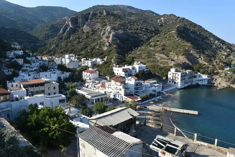 A small village of white flat-roofed houses stacked up a steep rocky slope above a narrow bay, with a short dark beach and a few parasols at the water's edge and a concrete jetty running out into turquoise water.