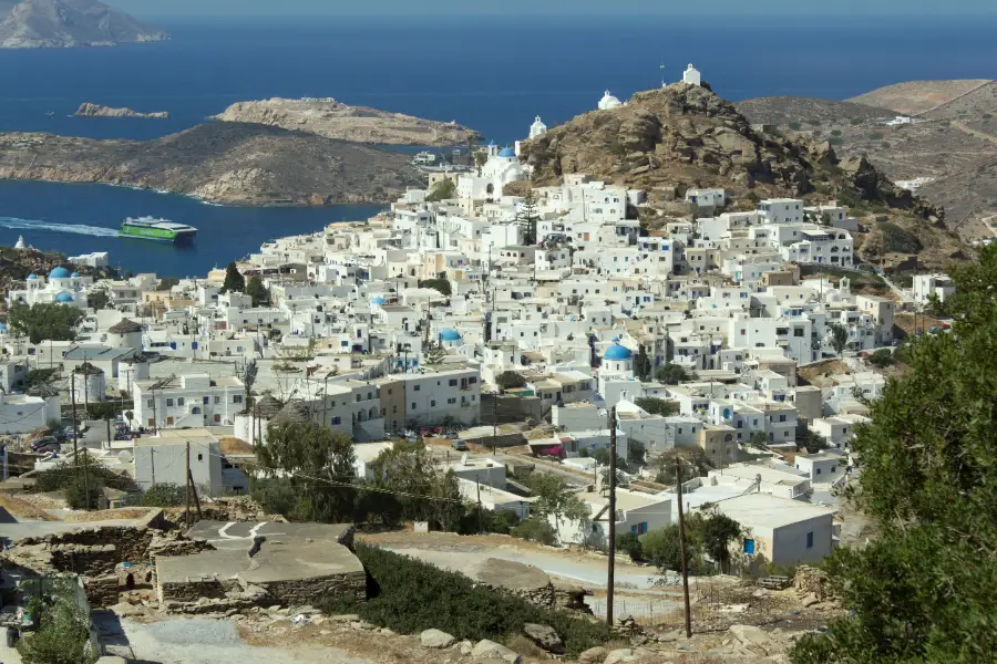 A Cycladic hill town of whitewashed cube houses stacked in tiers up a slope, with bare brown hills and the sea in the distance.