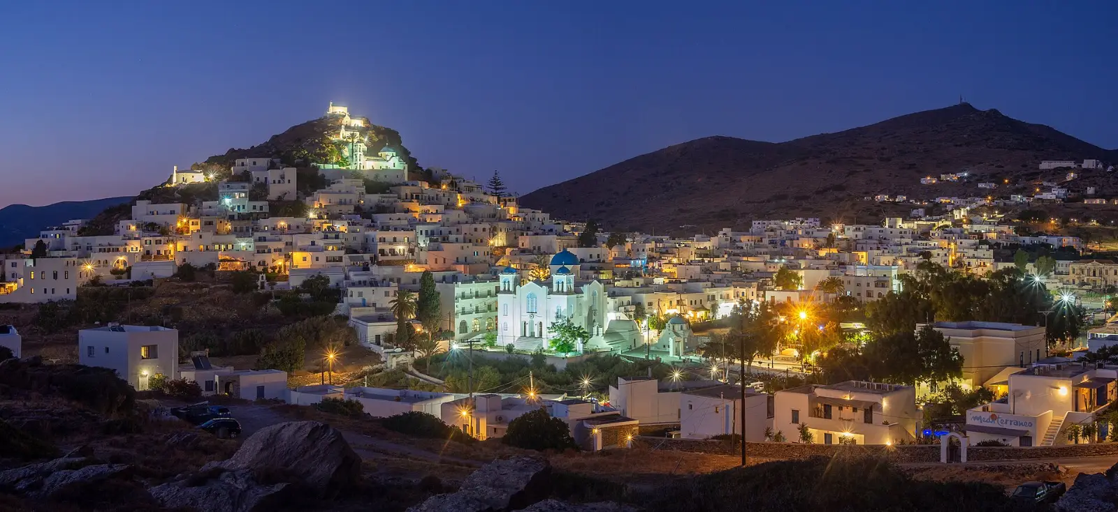 Ios Chora at blue hour, whitewashed cubic houses stacked up the hill with the summit chapel and a blue-domed church lit below
