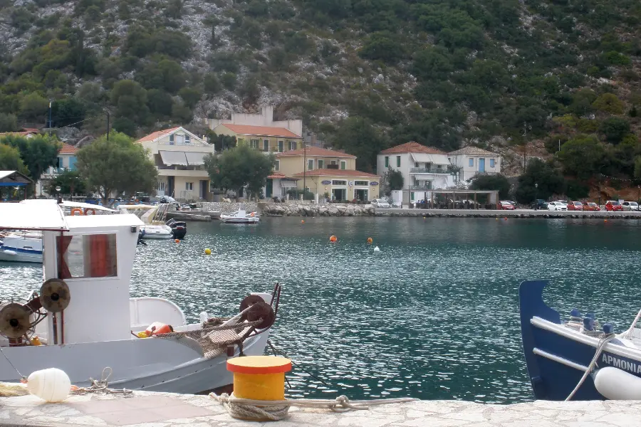 A small harbour with boats moored along a stone quay, low buildings behind it and steep green hillsides rising on both sides.