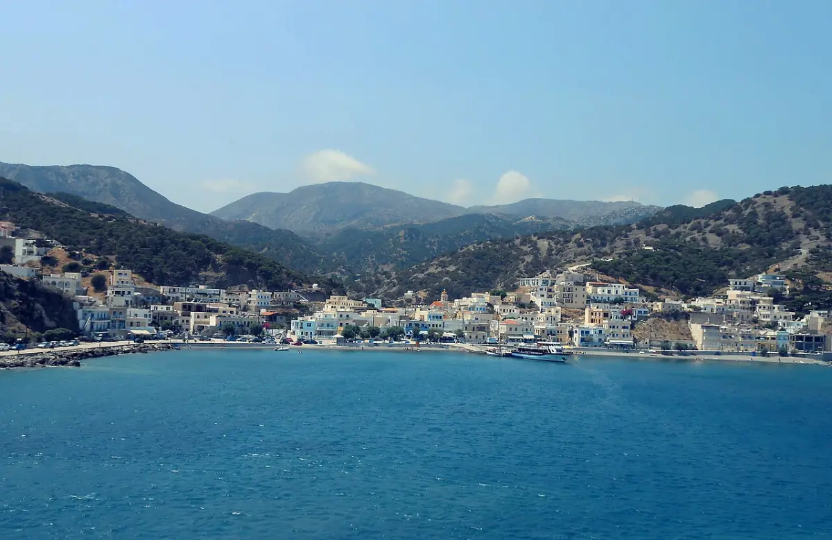 A small whitewashed coastal village spread along a bay at the foot of bare mountains, with an excursion boat moored at its quay and deep blue sea filling the foreground under a plain sky.