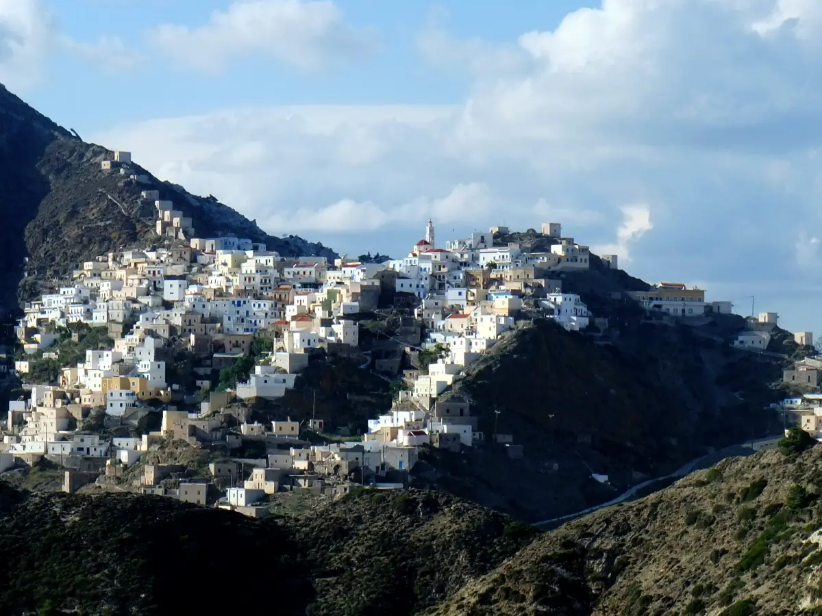 A village of flat-roofed white and ochre houses packed in tiers along a bare mountain ridge, with a church belfry near the high point and ruined stone buildings on the slope below.