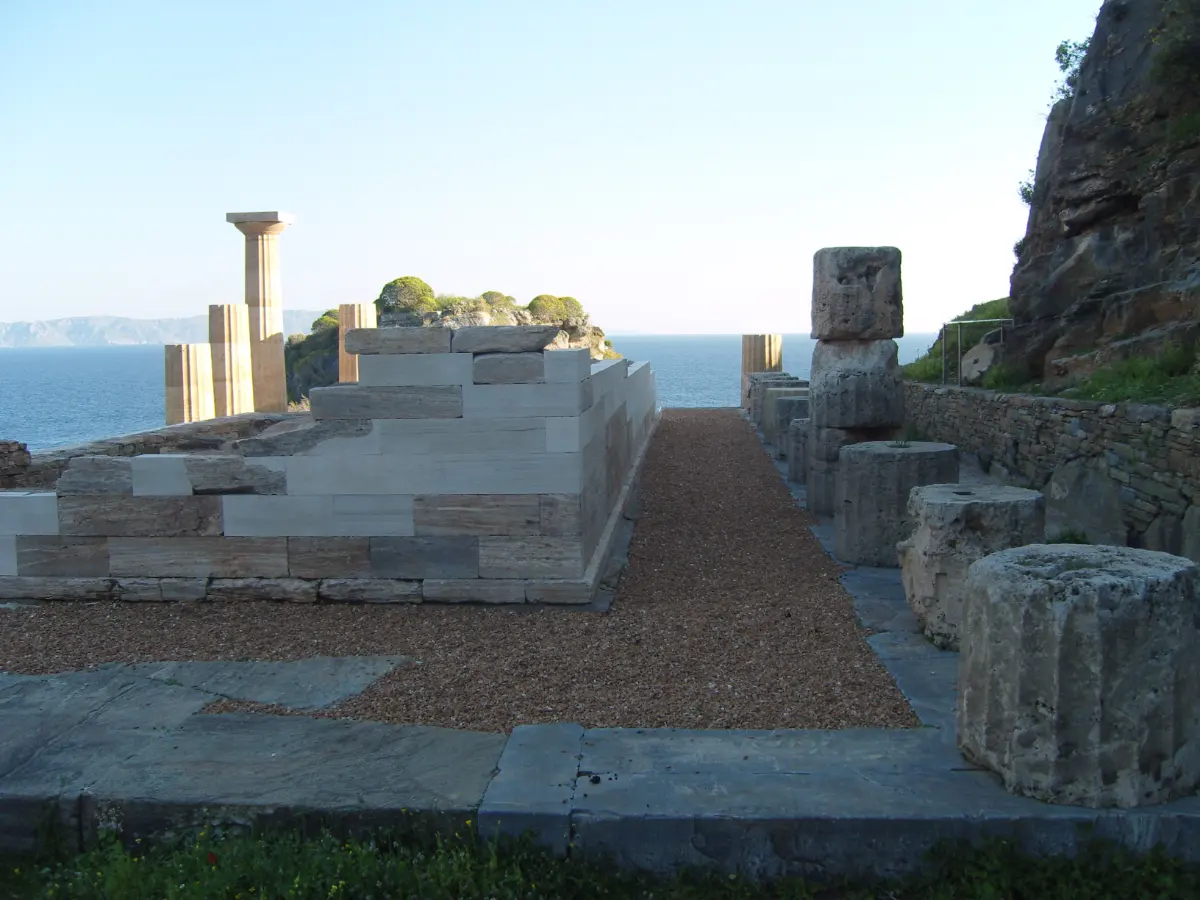 A temple platform of stone blocks on a terrace at the edge of the sea, with one part-rebuilt column standing at the far end and loose column drums lying on the ground beside it.