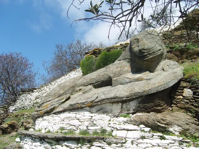 A weathered lion carved out of a single outcrop of grey bedrock on a terraced hillside, its head raised and its forelegs stretched out in front of it, with dry-stone walls and scrub around it.