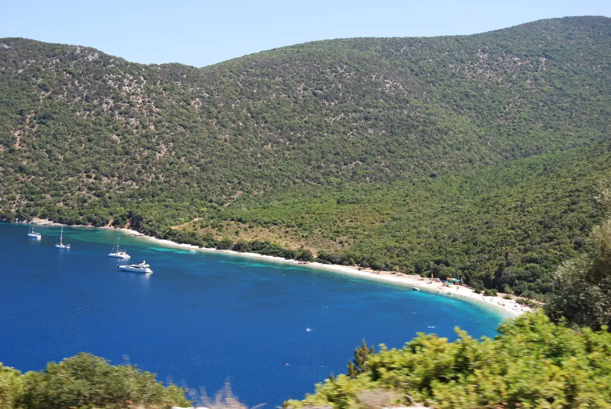 Antisamos bay on the east coast of Kefalonia: a long white pebble bank backed by steep, densely wooded green hills, with yachts anchored in clear deep blue water
