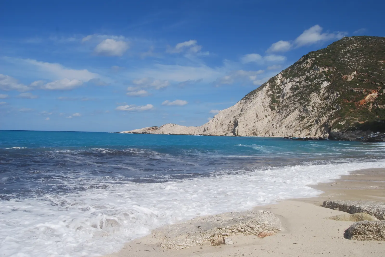 Petani beach on the Paliki peninsula of Kefalonia: surf breaking onto a pale pebble bank below a bare white limestone headland, with the sea stepping from turquoise to deep blue