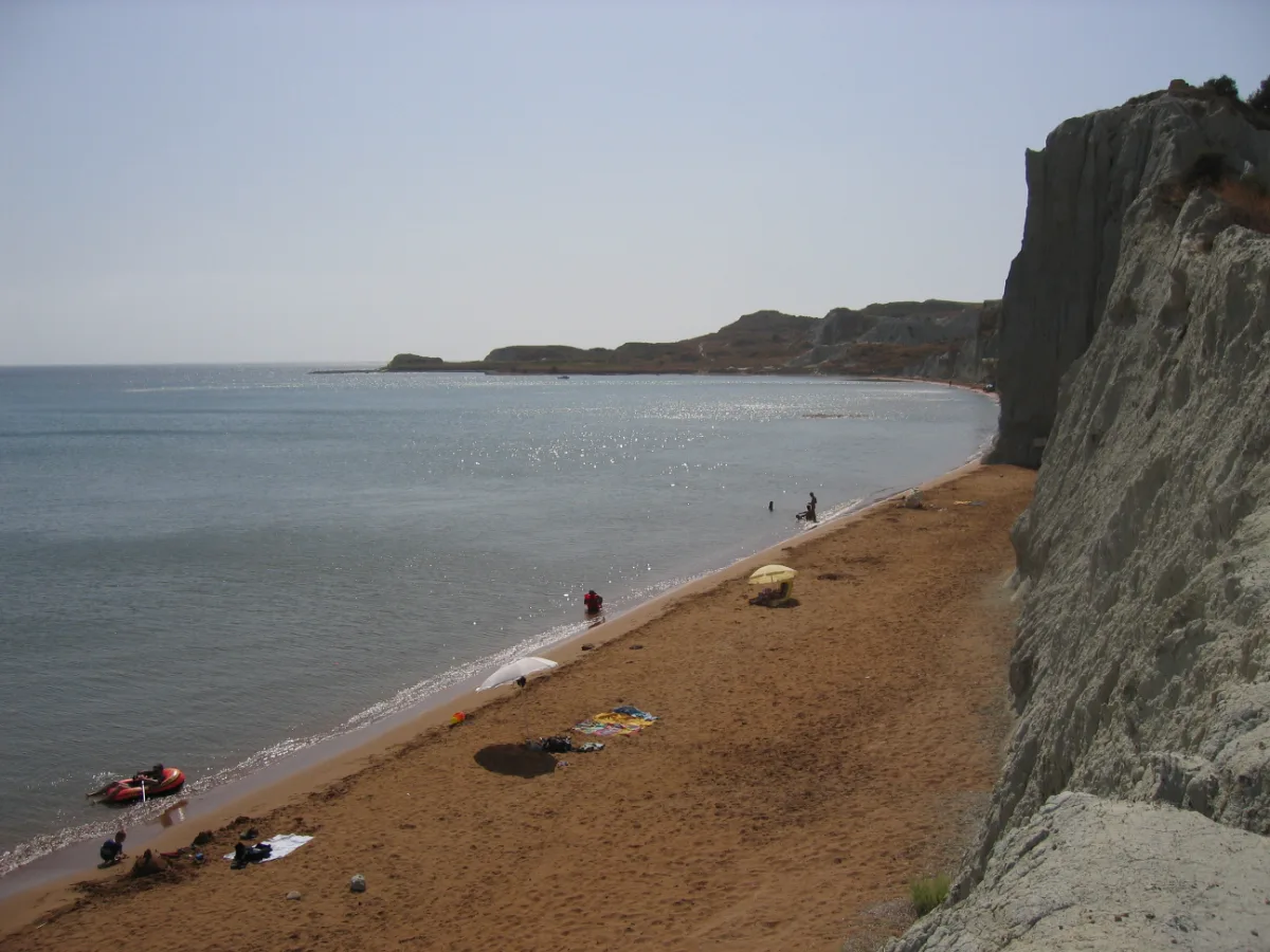 Xi beach on the Paliki peninsula of Kefalonia: a long curve of red-orange sand below cliffs of white clay, with shallow pale water in a calm bay