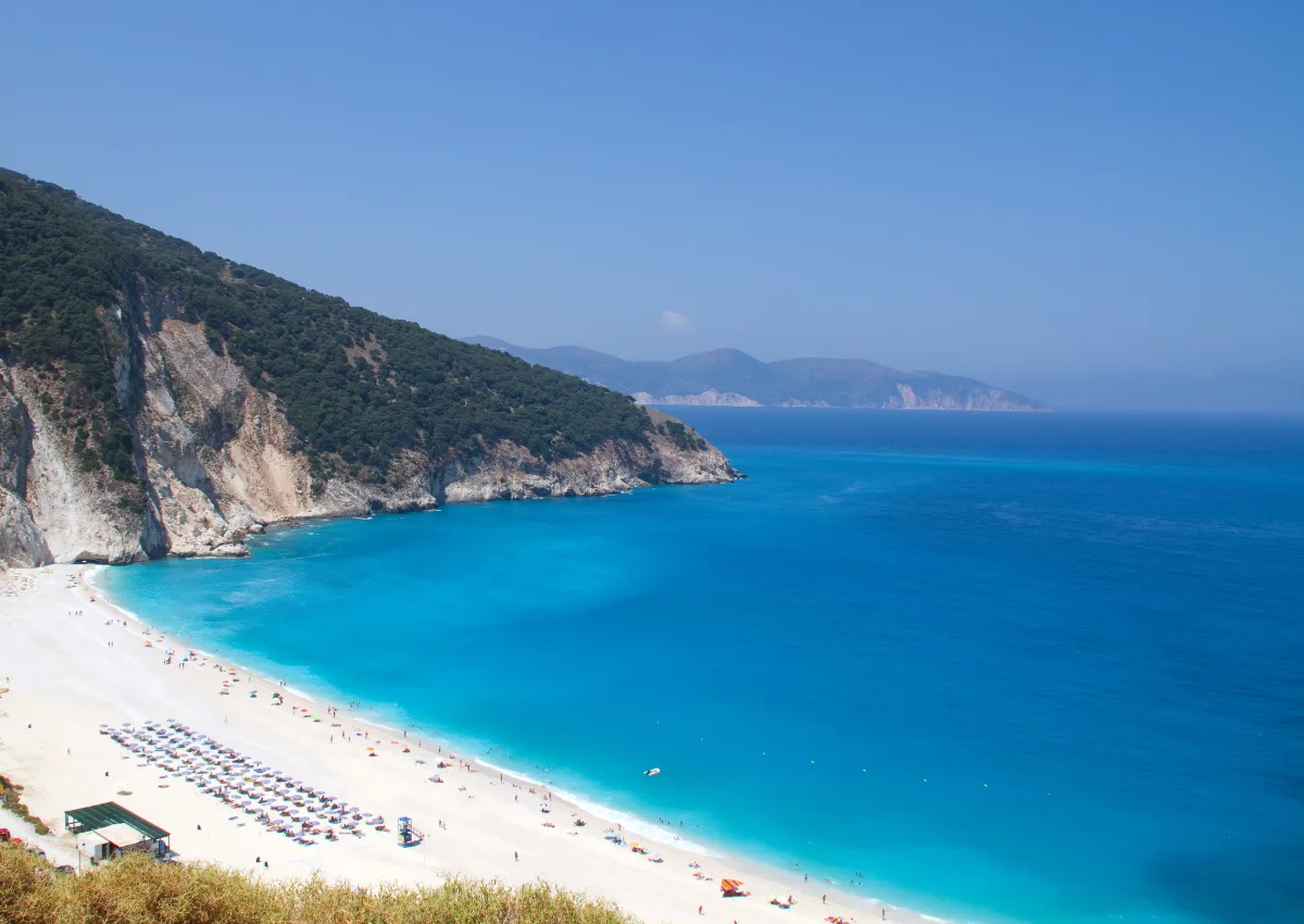 Myrtos beach in Kefalonia from high on the northern side: the white crescent with umbrella rows, a band of pale turquoise water stepping sharply to deep navy, and the limestone cliff with its access road cut across the face