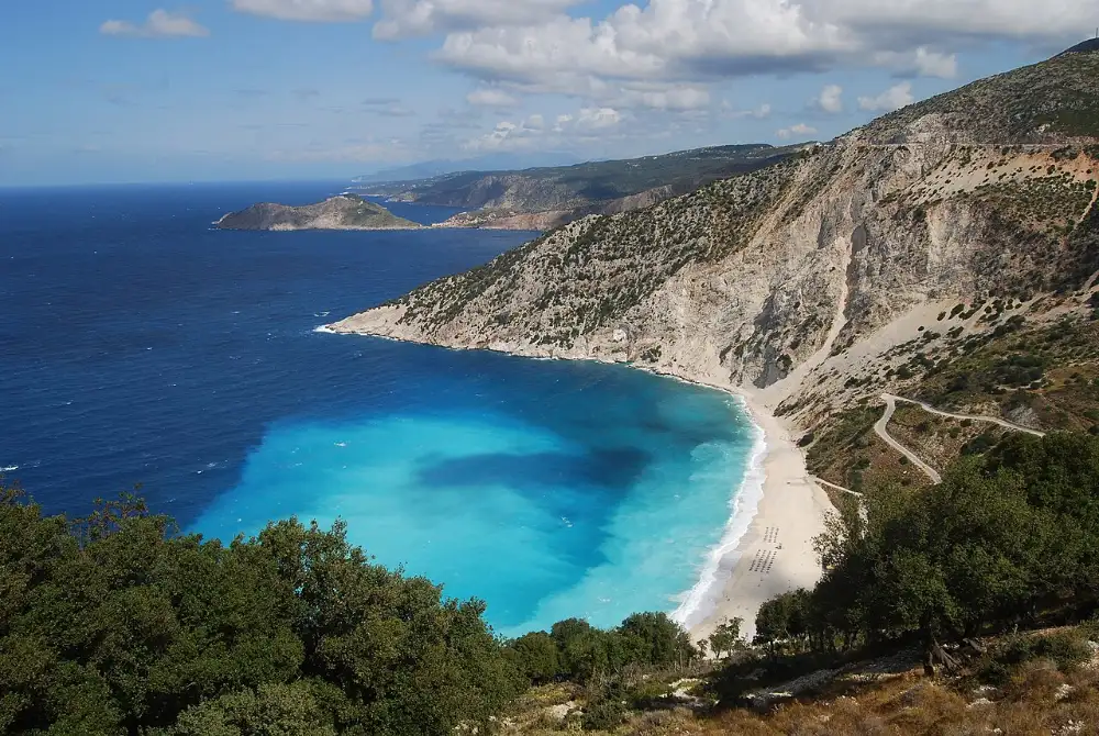 Myrtos beach seen from the road above, a white-pebble crescent under sheer cliffs with turquoise shallows and a navy bay