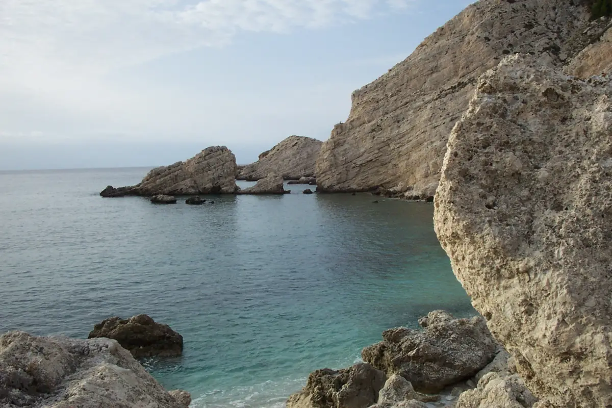 The white limestone cliff and low rock stacks closing the end of Petani bay on Kefalonia, dropping into glassy green water under an overcast sky