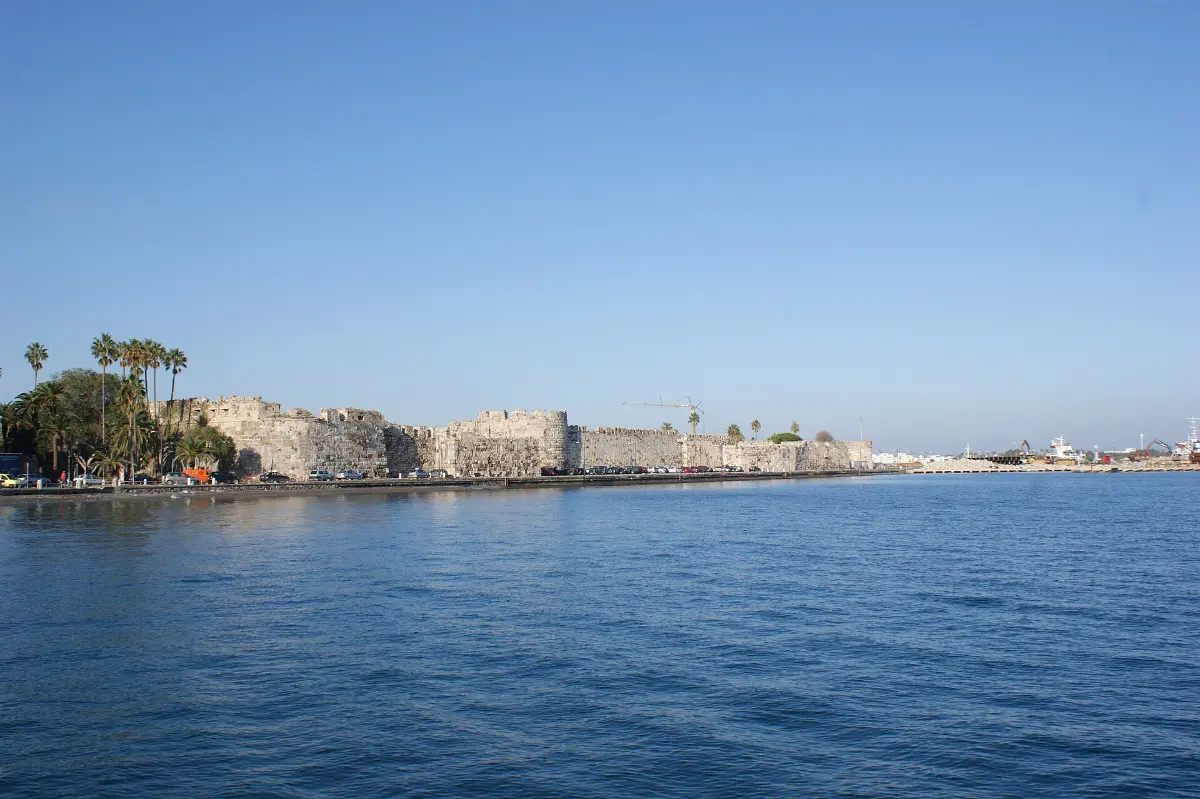 Neratzia Castle on Kos seen from the water: a long grey stone curtain wall with a square bastion above the harbour road, palm trees at one end, under a clear blue sky