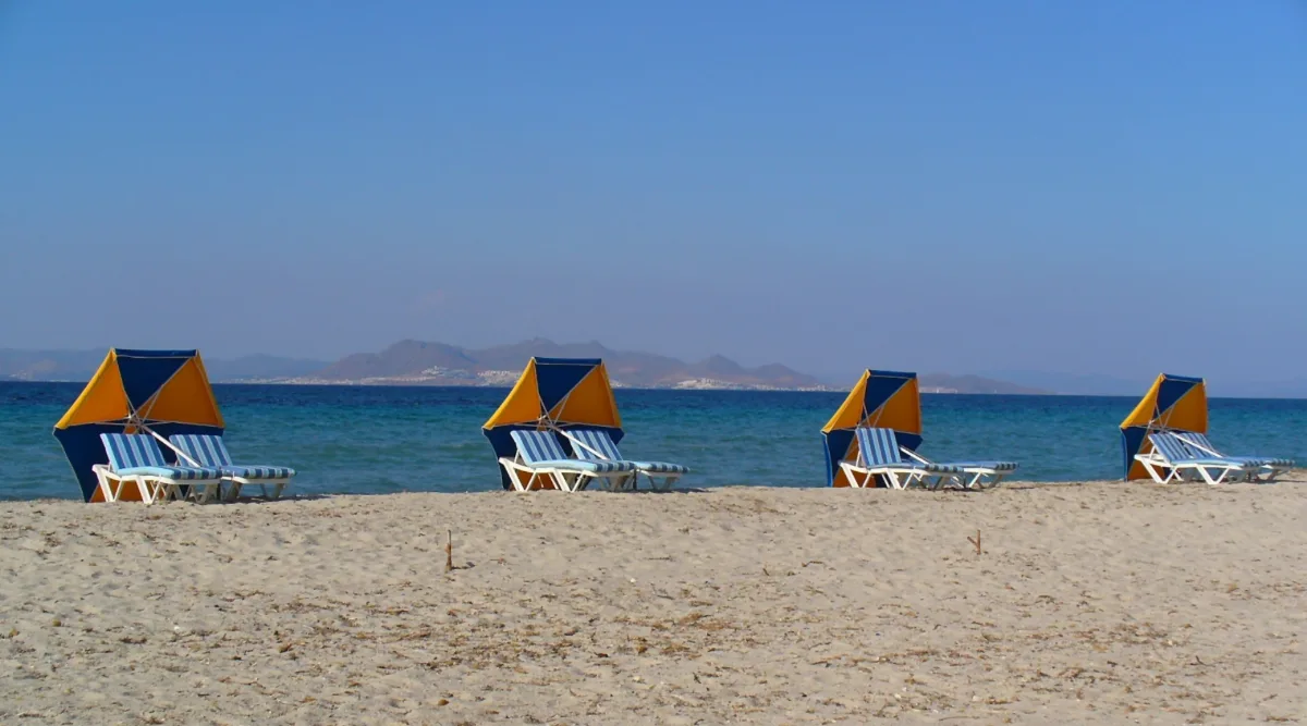 Empty sun loungers under blue and orange shades on the flat sand at Tigaki on Kos, with the Turkish coast across the water