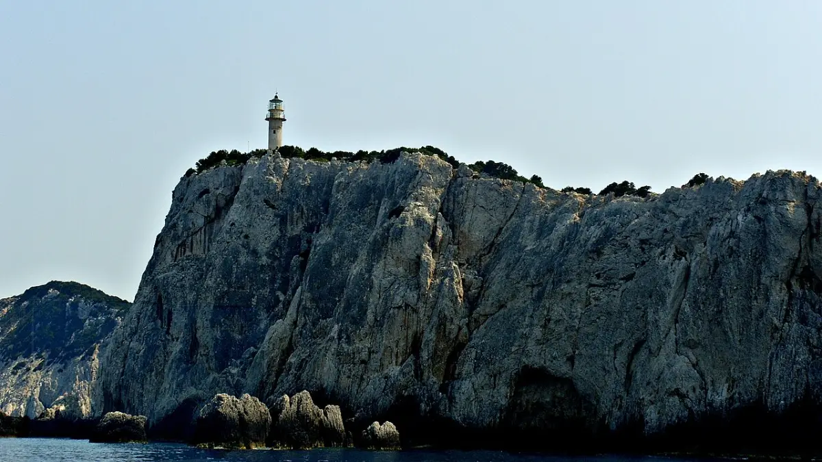 A white lighthouse tower standing on top of a massive pale limestone cliff that drops sheer to dark sea, under a plain hazy sky, with a lower rocky headland on the left and sea stacks at the base of the cliff.