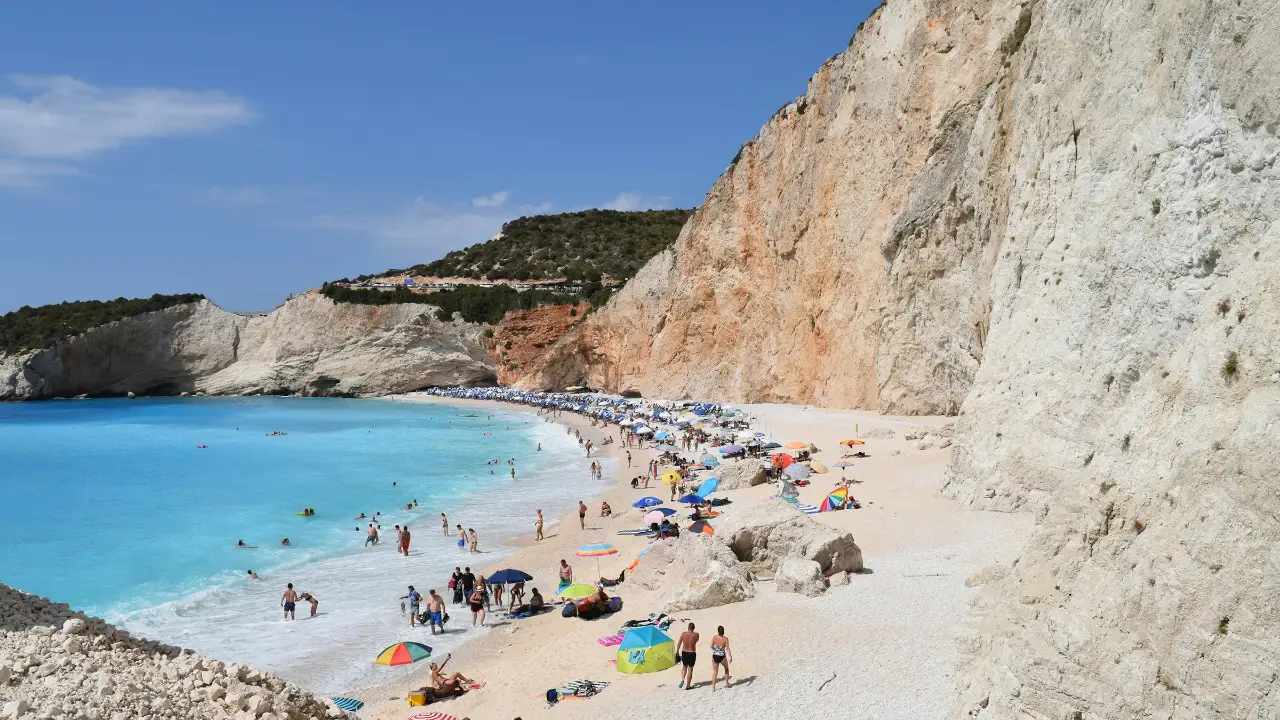 Porto Katsiki on Lefkada from the clifftop stair: the enormous white-and-ochre layered limestone cliff dropping vertically to a narrow white pebble beach, the milky-turquoise shallows shading to deep blue, and the green-topped headland running away to the left