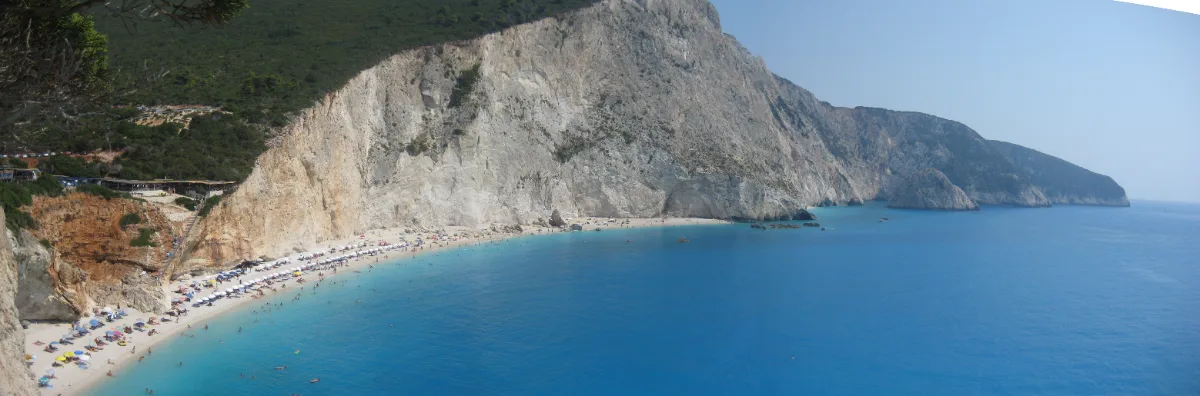 A curved strip of pale sand at the foot of a sheer white cliff, lined with rows of parasols, with deep turquoise water offshore and a headland of more white cliffs running away behind it.