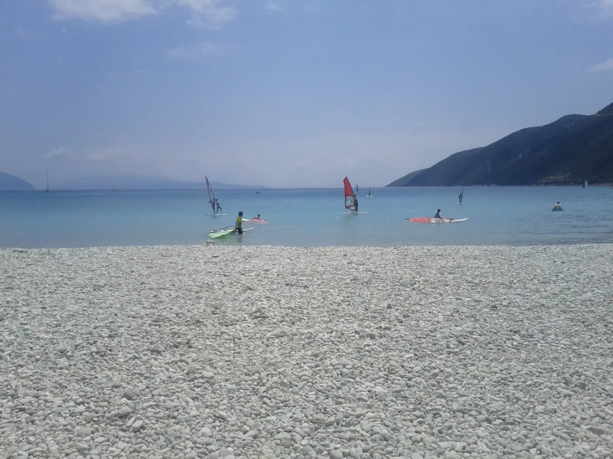A wide bank of white pebbles in the foreground and, beyond it, flat pale-green water where about a dozen windsurfers are sailing small rigs, with a green mountain ridge dropping to the sea on the right.