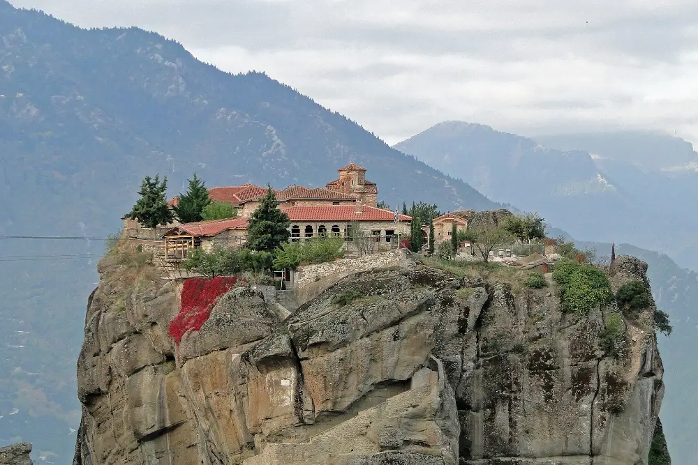The Monastery of the Holy Trinity at Meteora standing on top of a sheer sandstone pillar, with wooded mountain ridges fading into haze behind it