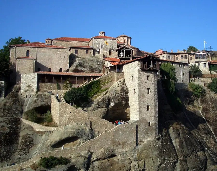 A large stone monastery on a flat rock summit: tiled roofs, a domed church, a tall square stone tower on the edge of the rock, and a walled staircase cut up the rock face with a few people climbing it.