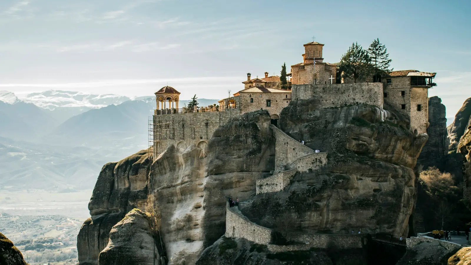 A Meteora monastery standing on the flat top of a sheer conglomerate rock pillar - stone cloister buildings, a domed katholikon and a small belfry-kiosk on the parapet, a stepped path and retaining walls cut into the rock below it, and the plain of Kalambaka with the Pindus range receding behind