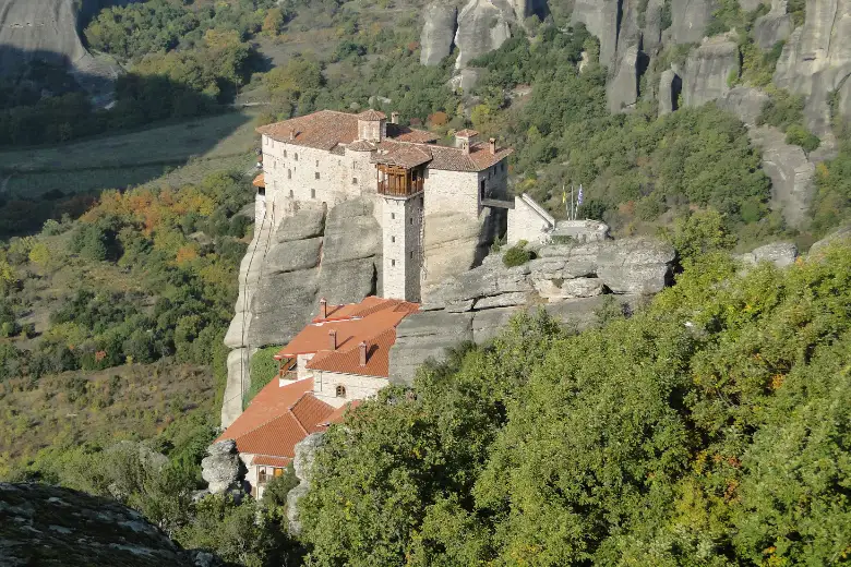 A monastery of stone walls and red-tiled roofs filling the whole flat top of a narrow rock pillar, its walls rising straight out of the rock, with wooded slopes and further grey pillars behind it.