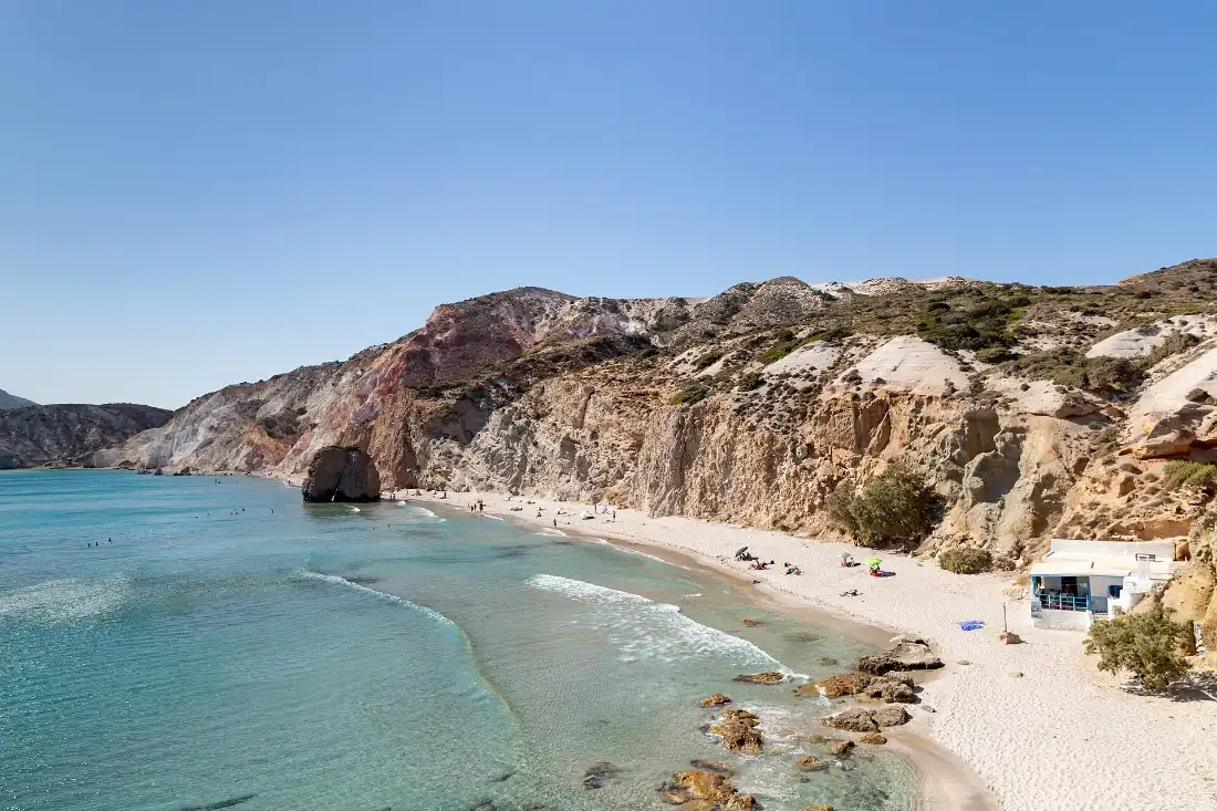 Firiplaka beach on the south coast of Milos: a curve of pale sand and turquoise water beneath banded white, red and ochre volcanic cliffs