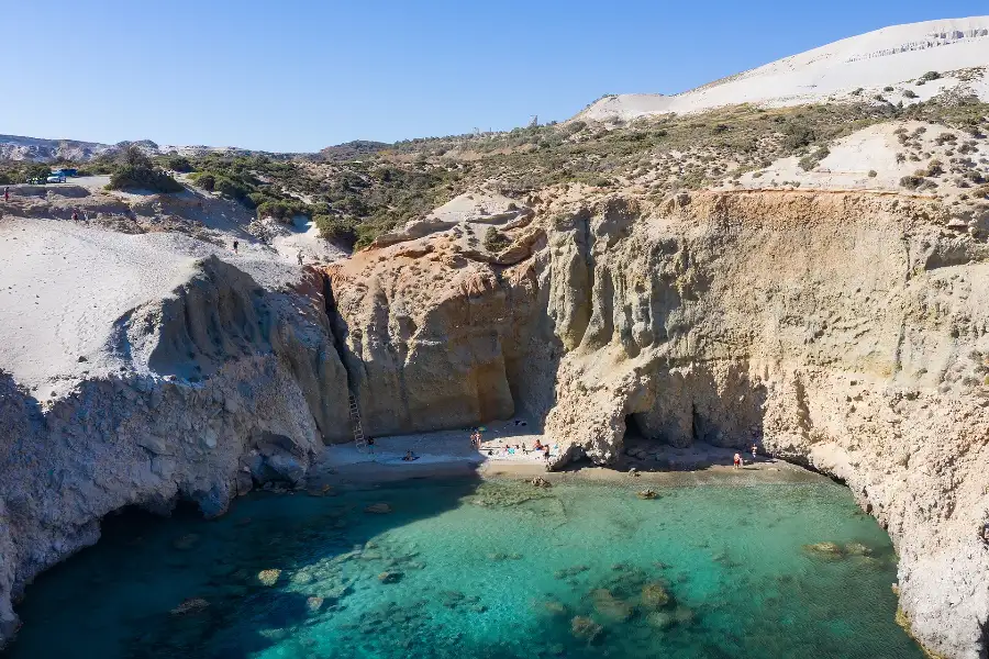 Tsigrado beach on Milos seen from the air: a small sand cove enclosed by cream and ochre cliffs, reached only by a wooden ladder in a narrow cleft in the rock